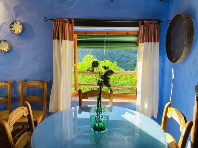 The image shows an indoor space, likely a dining area, with blue walls and a round glass table. There are wooden chairs arranged around the table, and a vase with plants is on the table. The window features white curtains and a view of greenery outside, suggesting a pleasant natural setting. Additionally, decorative plates hang on the wall, and a wooden basket is mounted nearby.